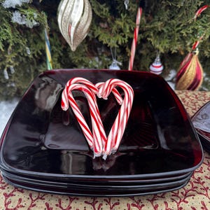 May include: A stack of four square, dark red plates with a glossy finish. Three red and white striped candy canes are arranged in a heart shape on top. The plates rest on a patterned tablecloth, with Christmas tree decorations in the background.