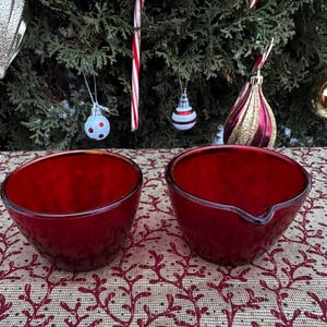 Könnte beinhalten: Zwei rubinrote Glasschalen mit strukturierten Oberflächen. Eine Schale hat einen Ausguss. Die Schalen stehen auf einer gemusterten Tischdecke, mit einem Weihnachtsbaum und Ornamenten im Hintergrund.