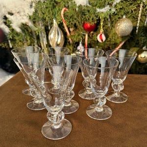 May include: A set of clear glass cordial glasses, each with a decorative bulbous stem and flared bowl. The glasses are arranged on a brown surface, with a Christmas tree and decorations in the background.