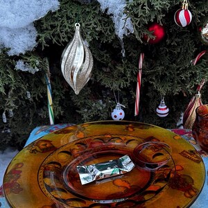 May include: A festive holiday scene featuring a decorated Christmas tree with ornaments, candy canes, and snow. A decorative amber-colored glass plate with a wrapped candy sits on a patterned tablecloth.