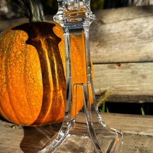 May include: A clear glass candlestick holder with a square base and a decorative top. An orange pumpkin is in the background. The candlestick holder is made of clear glass and has a rectangular shape.