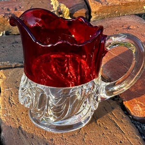 May include: A vintage glass pitcher with a ruby red upper half and a clear, ornate base. The handle is clear glass. The pitcher has a ruffled rim and is likely used for serving beverages. The pitcher is sitting on a brick surface.