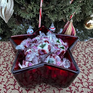 Puede incluir: Un plato de vidrio rojo en forma de estrella lleno de caramelos envueltos. Los caramelos son blancos con detalles rojos y verdes. El plato está sobre una superficie estampada, con un árbol de Navidad al fondo.