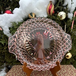 Puede incluir: Un plato de servir de cristal rosa con un patrón de diamantes y una sección central elevada. El plato se exhibe sobre un soporte de madera, con un fondo festivo de adornos y vegetación navideños.