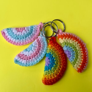 May include: Four semi-circular crochet keychains attached to silver metal rings. Two keychains feature pastel pink, white, and light blue yarn stripes. The other two keychains display vibrant rainbow stripes in red, orange, yellow, green, blue, and purple.