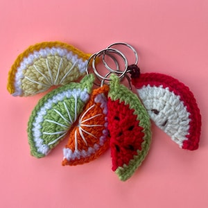 May include: A collection of five crocheted fruit slice keychains attached to silver metal rings. The set includes a yellow lemon slice, a green lime slice, an orange slice, a red watermelon slice, and a red and white apple slice, all featuring detailed yarn stitching.