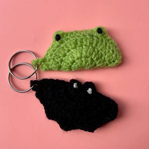 Könnte beinhalten: Zwei gehäkelte Schlüsselanhänger in Froschform. Ein Anhänger ist hellgrün mit Augen aus schwarzem Garn, der andere ist schwarz mit Augen aus weißem Garn. Beide sind an silberfarbenen Metallringen zur Befestigung von Schlüsseln angebracht.