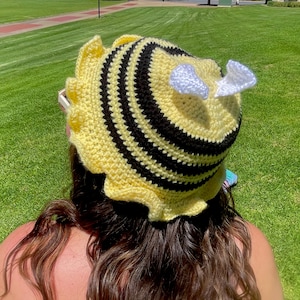 May include: A handmade crochet hat in a bee design. The hat is yellow with black stripes and white wings. The hat has a ruffled brim. The hat is designed to be worn on the head.