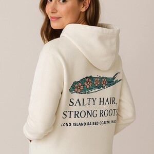 Op de afbeelding: Cr&egrave;mekleurige hoodie met een capuchon en lange mouwen. De achterkant heeft een teal en koraal bloemenontwerp over een Long Island-omtrek, met de tekst "SALTY HAIR, STRONG ROOTS" en "LONG ISLAND RAISED COASTAL MAD".