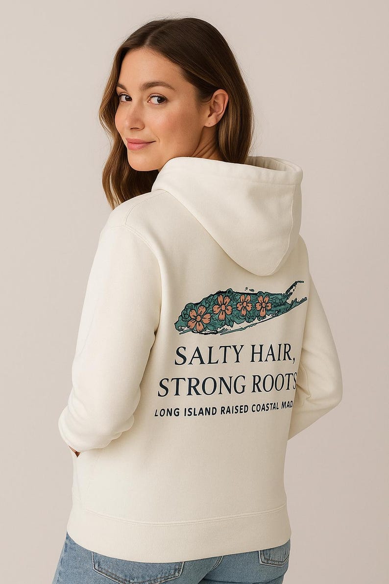 Op de afbeelding: Cr&egrave;mekleurige hoodie met een capuchon en lange mouwen. De achterkant heeft een teal en koraal bloemenontwerp over een Long Island-omtrek, met de tekst "SALTY HAIR, STRONG ROOTS" en "LONG ISLAND RAISED COASTAL MAD".