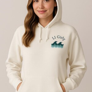 Op de afbeelding: Cr&egrave;mekleurige hoodie met een voorzak en een capuchon. De hoodie heeft de tekst "LI Girly" boven een afbeelding van oceaangolven in blauwgroen en blauw. Het model draagt een blauwe spijkerbroek.