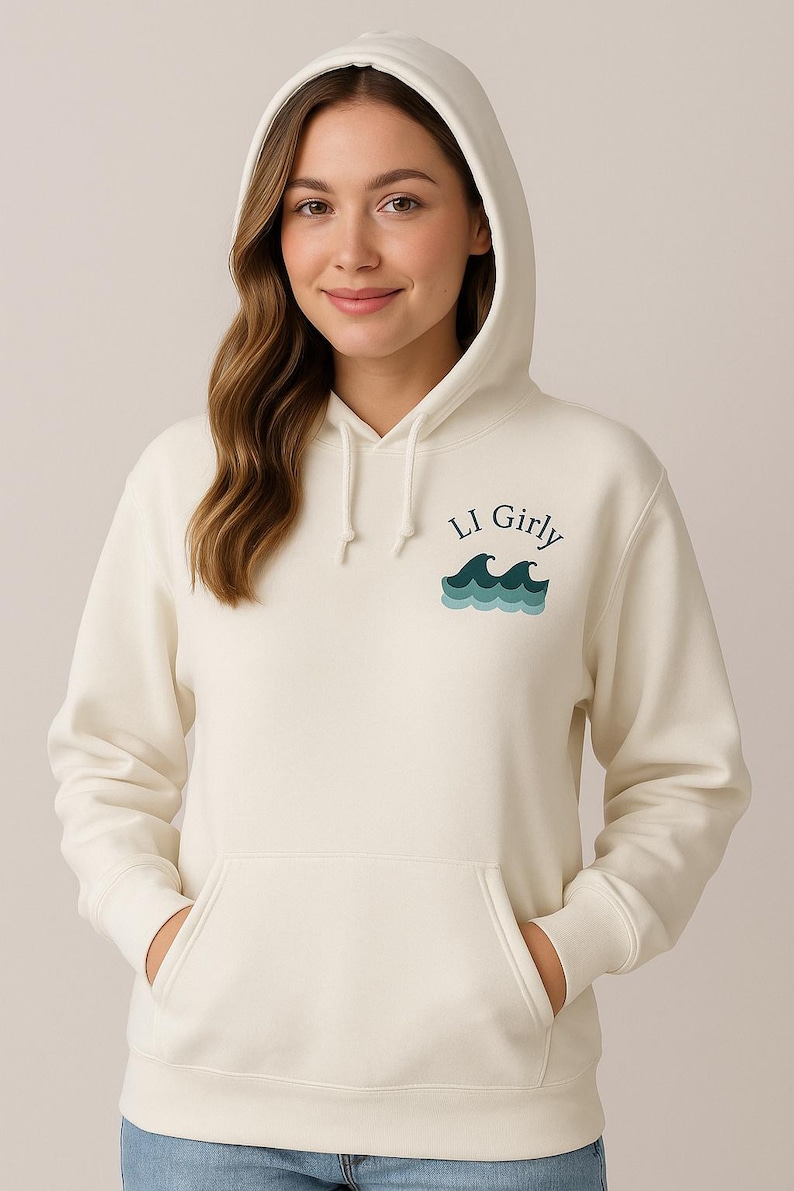 Op de afbeelding: Cr&egrave;mekleurige hoodie met een voorzak en een capuchon. De hoodie heeft de tekst "LI Girly" boven een afbeelding van oceaangolven in blauwgroen en blauw. Het model draagt een blauwe spijkerbroek.