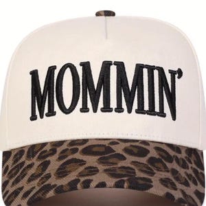 May include: A cream-coloured baseball cap embroidered with the word "MOMMIN'" in black. The peak has a brown and tan leopard print pattern. The cap features a curved peak and a button on top.