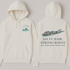 Op de afbeelding: Cr&egrave;mekleurige hoodie met een voorzak en capuchon. De voorkant heeft de tekst "LI Girly" boven een golfafbeelding. De achterkant toont een Long Island-omtrek met bloemaccenten en de tekst "Salty Hair, Strong Roots."