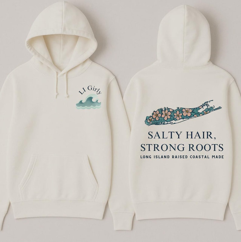 Op de afbeelding: Cr&egrave;mekleurige hoodie met een voorzak en capuchon. De voorkant heeft de tekst "LI Girly" boven een golfafbeelding. De achterkant toont een Long Island-omtrek met bloemaccenten en de tekst "Salty Hair, Strong Roots."