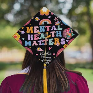 May include: Black graduation cap decorated with the words "Mental Health Matters" in colorful, retro-style lettering. The cap features rainbows, flowers, and the name "Sophia" with "University of Oxford 2024". A gold tassel hangs from the cap.