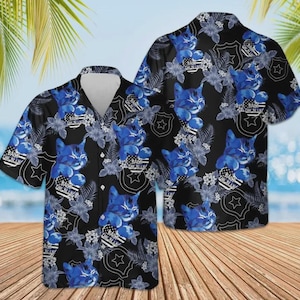 Puede incluir: Camisa negra de manga corta con un estampado de gato azul y flores. El diseño incluye un escudo con una estrella y el texto "Back the Blue". La camisa tiene cuello en V y cierre de botones.