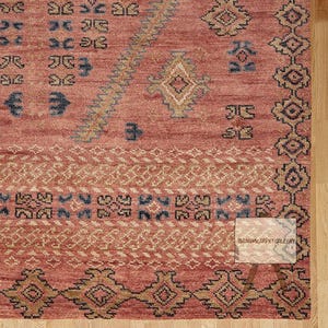 Peut inclure: Une section d'un tapis rouge avec des motifs géométriques complexes en beige, bleu et or. Le tapis présente un motif de bordure et un champ central avec des motifs répétitifs. Le texte "IBRAHIM CARPET GALLERY" est visible sur une petite étiquette.