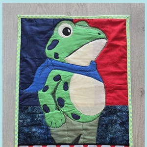 Puede incluir: Un tapiz de pared acolchado con un personaje de rana verde con manchas negras, que lleva una bufanda azul. El fondo está dividido en secciones rojas y azules. La palabra "RESIST" se muestra en la parte inferior, con frascos de insectos encima.