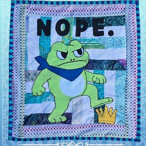 May include: A handmade quilt featuring a grumpy green frog wearing a blue bandana, stepping on a gold crown. The word "NOPE" is boldly displayed above the frog. The quilt has a patchwork background.