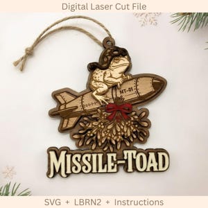 May include: A laser-cut wooden ornament featuring a toad wearing an aviator cap riding a missile labeled MT-01. The design includes a red bow, mistletoe, and the text MISSILE-TOAD at the bottom. A jute twine loop is attached for hanging.