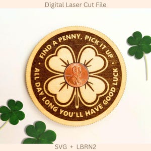 Op de afbeelding: Een ronde houten plaat met een lasergegraveerd ontwerp van een klavertjevier en een koperen penny in het midden. De tekst luidt: FIND A PENNY, PICK IT UP, ALL DAY LONG YOU'LL HAVE GOOD LUCK. Er zijn drie groene klaverbladeren zichtbaar.