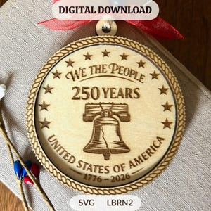 May include: A round wooden ornament with the text "We The People 250 Years United States of America 1776-2026" and a Liberty Bell illustration. The ornament has a rope-like border and is adorned with stars. A red ribbon is attached.