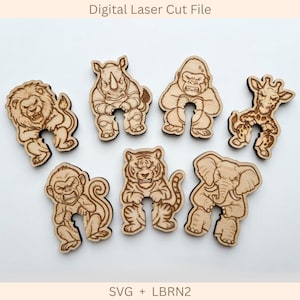 May include: A set of seven light-colored wooden animal cutouts featuring a lion, rhinoceros, gorilla, giraffe, monkey, tiger, and elephant. Each figure has a U-shaped cutout at the bottom. The text Digital Laser Cut File and SVG + LBRN2 is visible.