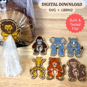 Op de afbeelding: Een set houten broodclips in dierenvormen, waaronder een leeuw, gorilla, olifant, neushoorn, giraf, tijger en aap. De tekst luidt DIGITAL DOWNLOAD SVG + LBRN2 en Built & Tested File! Eén leeuwenclip is bevestigd aan een plastic broodzak.