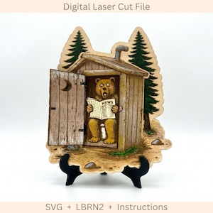 May include: A digital laser cut file featuring a wooden outhouse with an open door showing a brown bear sitting inside reading a newspaper labeled NEWS. The scene includes two green pine trees, a crescent moon cutout on the door, and a pipe.