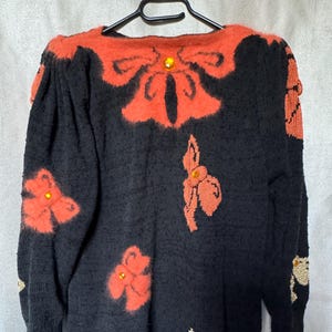 Peut inclure: Pull noir avec des nœuds et des motifs floraux orange. Le pull a un col rond et des manches longues. Les détails de nœuds et de fleurs sont ornés de pierres précieuses jaunes.