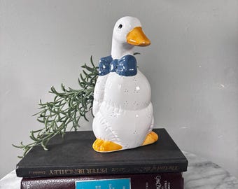 May include: A white ceramic goose figurine featuring a glossy finish, a bright orange beak and feet, and a dark blue bowtie. The goose has small black dots patterned across its body and is positioned standing upright with a smooth, rounded shape.