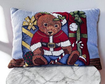 May include: A rectangular Christmas-themed pillow featuring a brown teddy bear dressed in a Santa Claus outfit. The bear is surrounded by wrapped gifts and a candy cane. The pillow has a light blue background and is made of woven fabric.