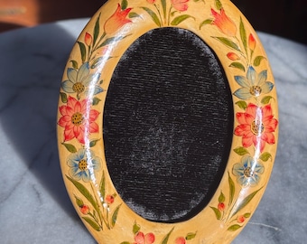 May include: Oval-shaped, hand-painted picture frame with a cream-colored base. The frame is adorned with red, blue, and orange floral designs and green leaves. The center has a black velvet backing. A decorative item for displaying photos.