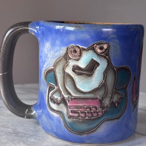 Könnte beinhalten: Blaue Keramik-Tasse mit schwarzem Henkel. Die Tasse zeigt ein skurriles Froschdesign in Blau-, Türkis- und Rosatönen. Der Frosch sitzt auf einem Seerosenblatt. Die Tasse hat eine glänzende Oberfläche.