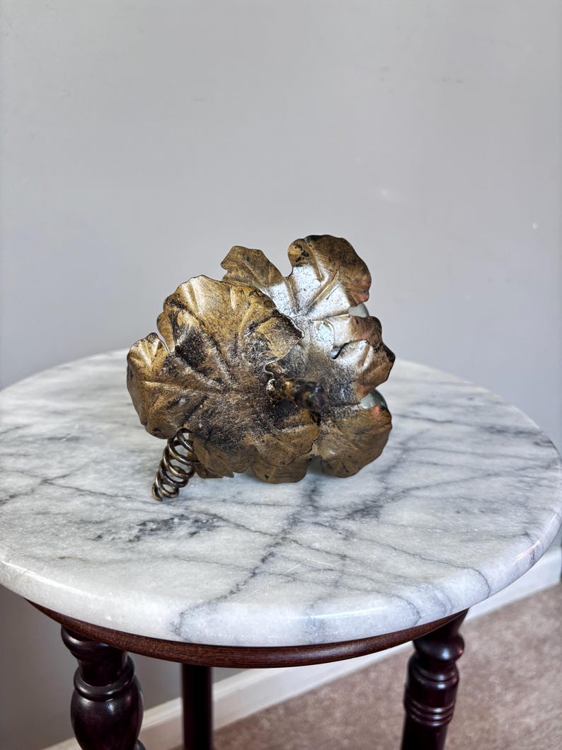 May include: A decorative metal sculpture shaped like a cluster of grapes with large, textured leaves finished in a distressed antique gold tone. The piece features a coiled wire stem extending from the base of the metallic foliage cluster.