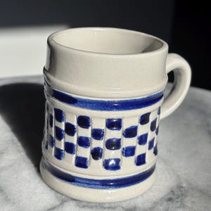 May include: A ceramic mug with a cream-colored base and a blue checkered pattern. The mug has a handle and two blue stripes. The mug is cylindrical and appears to be handmade.