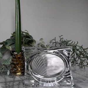 May include: Clear glass dish with an oval center and a square, textured frame. The dish sits on a marble surface, with a green candle and gold pineapple candle holder to the left. Greenery is visible in the background.