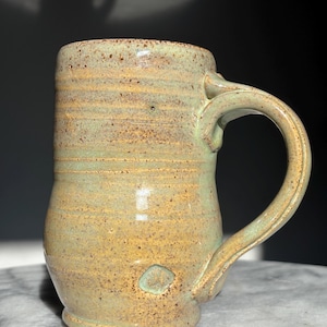 May include: Handmade ceramic mug with a speckled, earthy green and tan glaze. The mug features a large handle and a slightly flared shape. The stoneware mug has a rustic, artisanal aesthetic, perfect for coffee or tea.