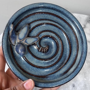 May include: A round ceramic dish with a glossy blue glaze and a dark brown spiral pattern etched into the center. A small, three-dimensional sculpted olive branch with three leaves and one olive is attached to the inner rim of the dish.