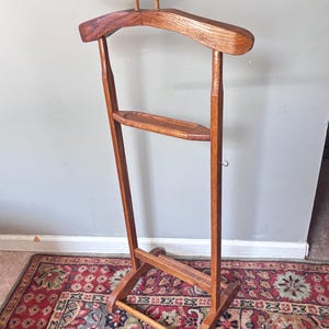 May include: A freestanding wooden valet stand featuring a wide contoured hanger at the top for jackets, a straight horizontal bar above it for accessories, a small central shelf for personal items, and a lower horizontal bar for trousers.