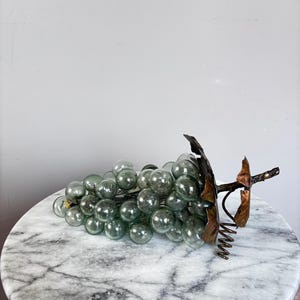 May include: A decorative cluster of translucent, pale green glass spheres arranged to resemble a bunch of grapes. The cluster features a dark, metallic stem and textured, bronze-colored leaves attached to the top, with a coiled wire tendril.