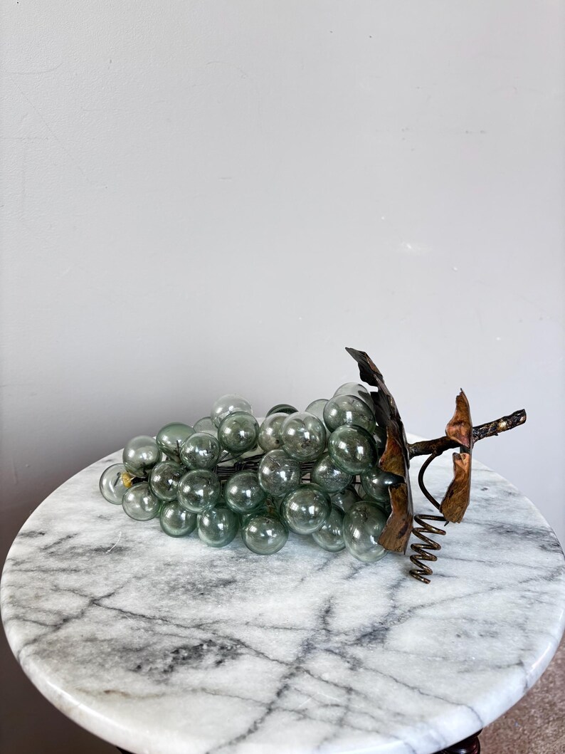 May include: A decorative cluster of translucent, pale green glass spheres arranged to resemble a bunch of grapes. The cluster features a dark, metallic stem and textured, bronze-colored leaves attached to the top, with a coiled wire tendril.