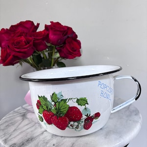 May include: A white enamel bowl with a black rim and a single handle, featuring a speckled finish and a graphic of red strawberries with green leaves. The text "POPCORN BOWL" is printed in blue on the side of the bowl in a casual font.