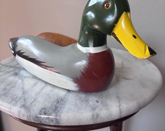 May include: A hand-painted wooden duck figurine with a green head, yellow beak, and gray body, resting on a marble-topped side table. The duck has a white stripe around its neck and a red breast. The table has dark brown legs.