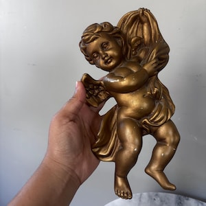 May include: A gold-colored wall plaque featuring a cherub with wings, depicted in a flying pose while holding a draped fabric. The figure has detailed facial features and curly hair, with a metallic finish and visible signs of wear.
