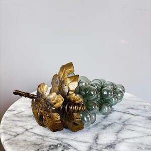 May include: A decorative cluster of translucent, pale green glass grapes attached to a textured, metallic gold-colored stem featuring two large, detailed leaves and a small coiled wire accent extending from the base of the foliage.