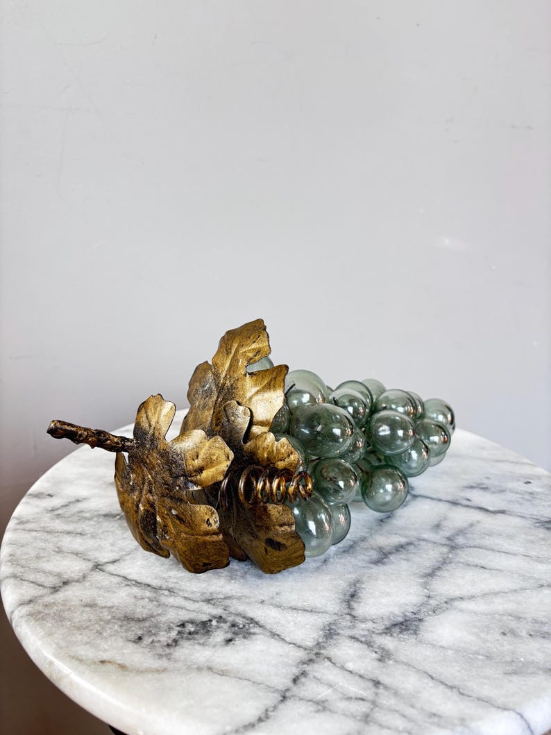 May include: A decorative cluster of translucent, pale green glass grapes attached to a textured, metallic gold-colored stem featuring two large, detailed leaves and a small coiled wire accent extending from the base of the foliage.