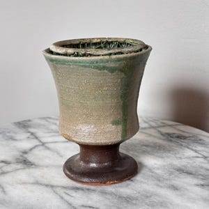 Puede incluir: Un cáliz de cerámica con borde acampanado y base de pedestal. La parte superior presenta un acabado beige texturizado y moteado con un goteo de esmalte verde, mientras que la base es de arcilla lisa de color marrón oscuro.