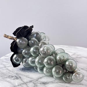 May include: A decorative cluster of translucent, pale green glass grapes attached to a dark, textured metal stem and leaf. The individual glass spheres are rounded and smooth, creating a realistic bunch of fruit with a metallic accent at the top.